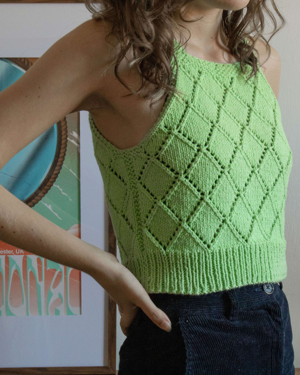 Close up of the front of a person wearing a green knitted vest top in front of a white wall and a framed print