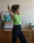 Person wearing a green knitted vest top and dark jeans standing in a room with wooden furniture and plants.