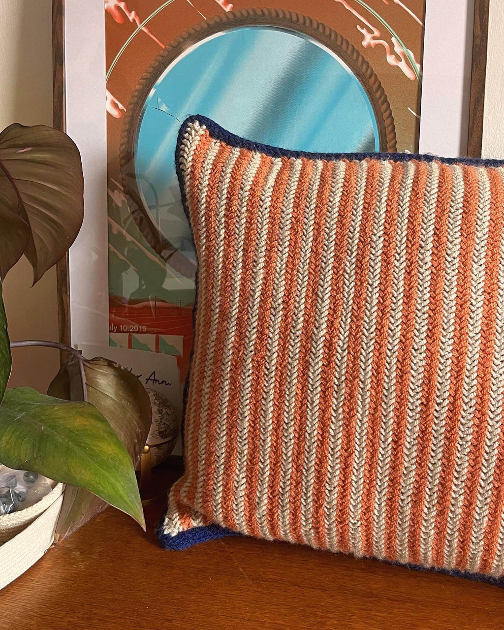 Striped herringbone stitch pillow on a wooden surface with a decorative print and plant in the background